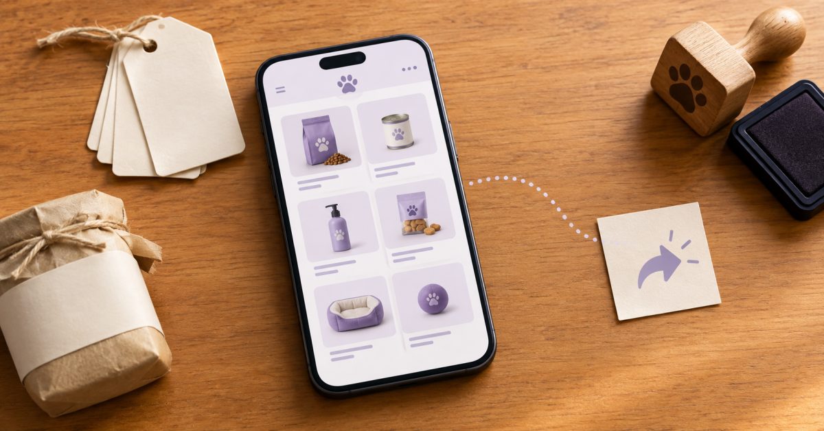 Celular mostrando grade de produtos de um catálogo digital, etiquetas em branco, embrulho de produto, carimbo de patinha e seta de compartilhar sobre mesa de madeira